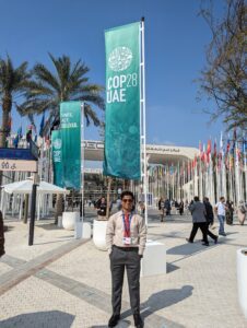 Vinay Karthik in a beige shirt, red tie, and grey pants standing in front of the COP28 banner. Palm trees and flags of countries around the world are in the background.