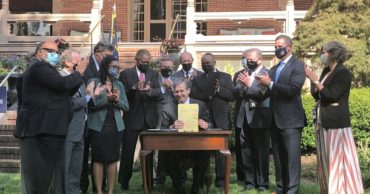 Photo of Gov. Cooper holding up signed bill with members of bipartisan legislature clapping and smiling around him. Gov. is seated others are standing.