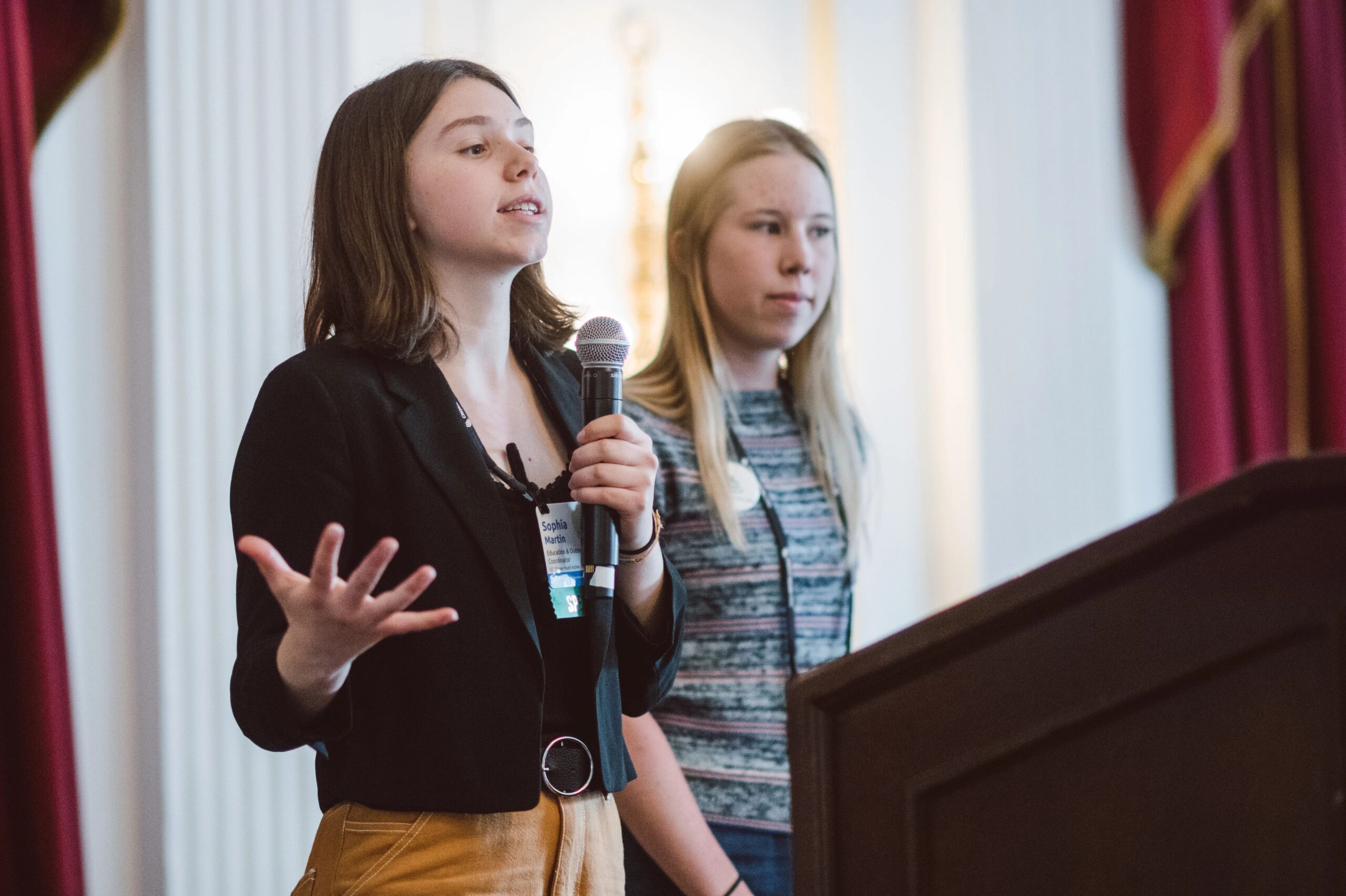 Two members of the Tahoe Youth Action Team are talking about their school electrification campaign on stage. The person on the left is holding a microphone and speaking. Behind them is a window with red curtains.